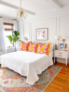 a white bed with colorful pillows and blankets on it in a bedroom next to a window