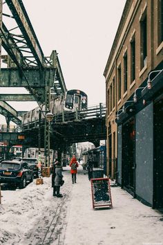 two people walking down a snowy street next to tall buildings and a train on the tracks