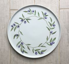 a white plate with blue berries and green leaves painted on the rim is sitting on a tile floor