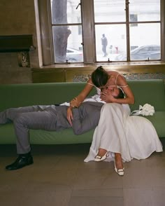 a bride and groom sitting on a green couch in front of large windows, kissing