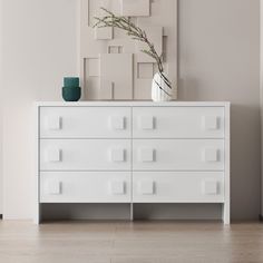 a white dresser sitting next to a vase with a plant in it and a painting on the wall