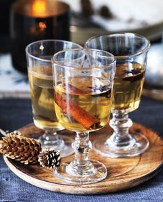 three glasses filled with different types of alcohol on a wooden tray next to a pine cone