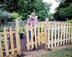 a woman standing next to a wooden fence in a garden with plants and flowers around her