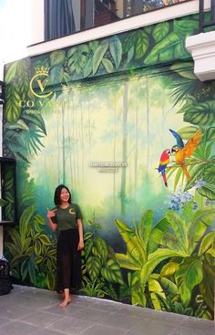 a woman standing in front of a mural on the side of a building with birds and trees