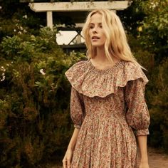 a woman in a floral dress is standing near some bushes and trees, looking off to the side