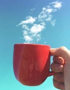 a hand holding a red coffee cup in front of a blue sky with white clouds