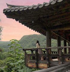 a woman standing on top of a wooden balcony next to a lush green forest covered hillside