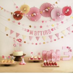 a table topped with pink and gold cupcakes next to paper fan garlands