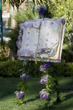 an open book decorated with purple flowers and greenery in front of a garden area