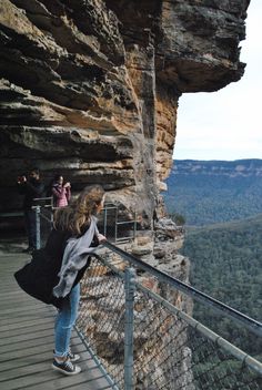 people standing at the edge of a cliff looking out over trees and mountains from a viewing platform