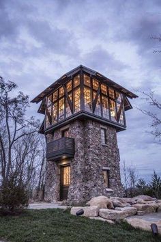 a stone tower with glass windows on the top is lit up at night in front of trees and rocks