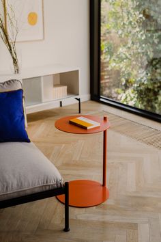 an orange table sitting in front of a window next to a couch with a book on it