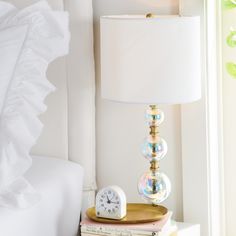 a nightstand with a lamp, books and a clock on it next to a window