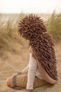 a young child sitting in the sand wearing a hat made out of leaves and feathers