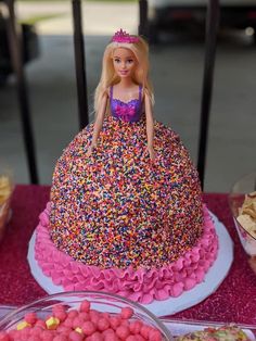 a barbie doll cake with sprinkles on it and other desserts around it