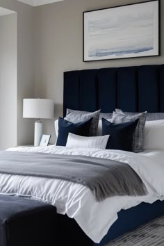 a bed with white and blue pillows in a bedroom next to a window, framed painting on the wall