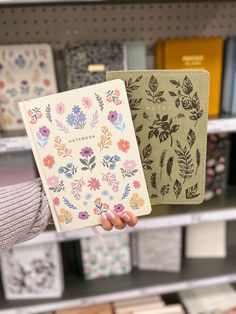 a person holding two notebooks in front of a book shelf with books on it