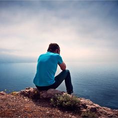 a man sitting on top of a rock next to the ocean looking out at the water