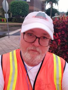 a man with glasses and a white beard wearing an orange vest is standing in front of some bushes