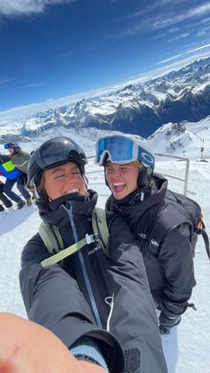 two people taking a selfie on the top of a ski slope with snow capped mountains in the background