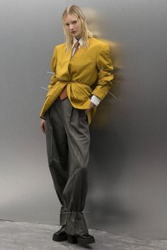 a woman standing in front of a gray wall wearing a yellow jacket and grey pants