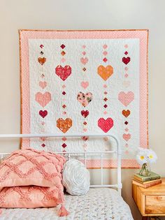 a white bed topped with a pink and orange quilt next to a wooden nightstand filled with flowers