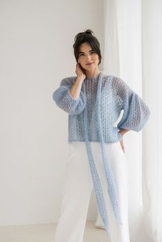 a woman standing in front of a window wearing a blue knitted sweater and white pants