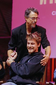 two men sitting in chairs with one being hugged by the other