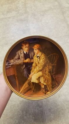 a person holding up a plate with a painting on it's side that has two men sitting at a table