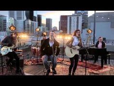 three women singing and playing guitar on a roof top with city lights in the background