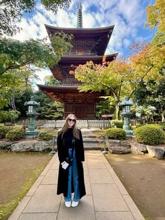 a woman standing in front of a tall pagoda with trees and bushes around her feet