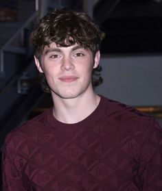 a young man with curly hair wearing a maroon shirt and smiling at the camera while standing in front of some stairs