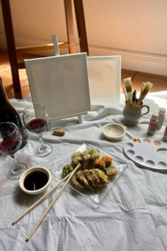 a table topped with plates and bowls filled with food next to wine glasses, paintbrushes and an easel