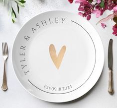 a white plate with a gold heart on it next to silverware and pink flowers