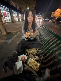 a woman sitting on a bench eating french fries and hotdogs in front of her