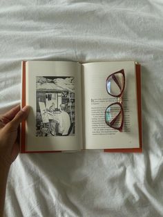 an open book with glasses on it laying on top of a white bed next to a pair of reading glasses