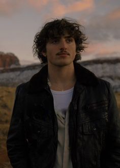 a man with long hair wearing a black jacket and standing in front of a mountain