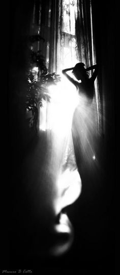 a black and white photo of a woman standing in front of a window with the sun shining through