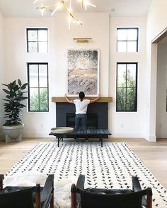 a man sitting on a black and white rug in front of a fire place with two large windows