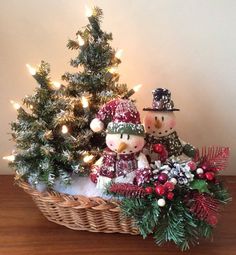 two snowmen sitting in a basket next to a christmas tree with lights on it