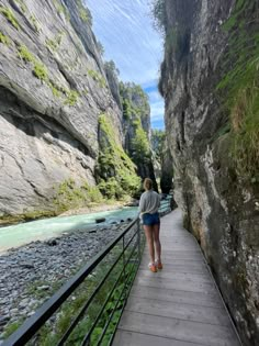 a woman walking down a wooden walkway next to a river in a canyon with green moss growing on the rocks