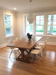 a dining room table with white chairs in front of two windows and a potted plant