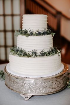 a three tiered white cake with greenery on top sits on a silver platter