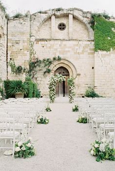 an outdoor ceremony setup with white chairs and greenery on the wall, in front of a stone building