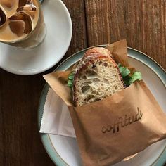 a sandwich in a paper bag sitting on top of a plate next to a cup of coffee