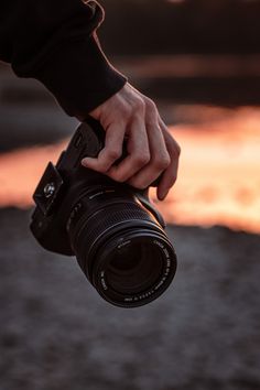 a person holding a camera in their hand with the sun setting behind them and reflecting on the water