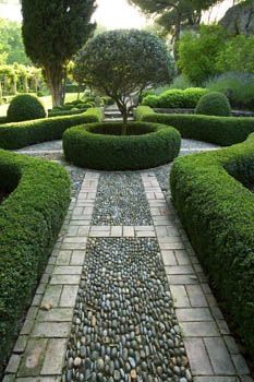 a stone path in the middle of a garden surrounded by hedges and trees, leading to a small tree