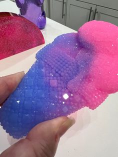 a hand holding a piece of soap on top of a white table next to two purple and pink objects