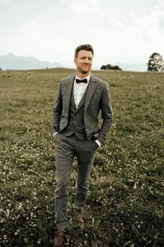 a man standing in a field wearing a suit and bow tie with his hands in his pockets