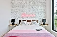 a bed with pink and white sheets in a bedroom next to a large window that has a neon sign above it